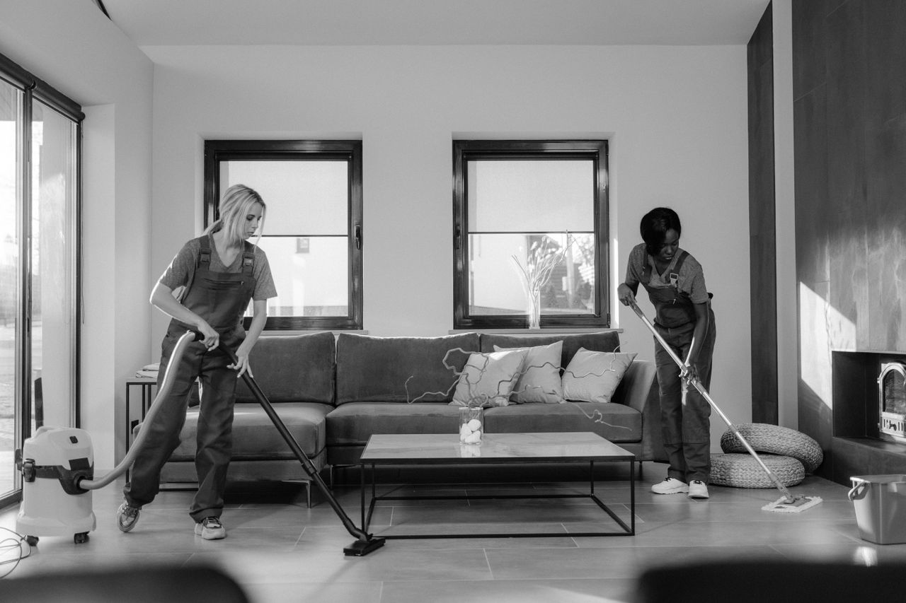 Two women dressed for work cleaning a contemporary living room with a vacuum and a mop.